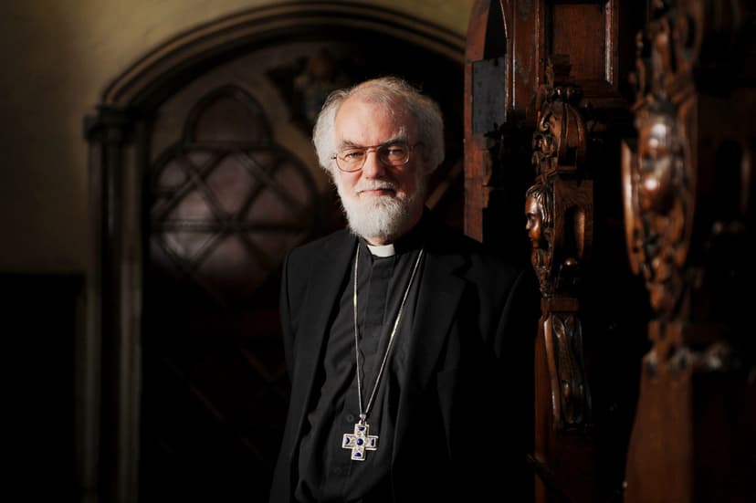 Rowan Williams with cross around neck standing in front of a large oak door